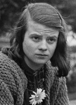 Black and white photo of a young woman with shoulder-length hair, looking pensive. She's wearing a knitted sweater and has a small flower pinned to her clothing. The background is blurred, focusing on her expression.