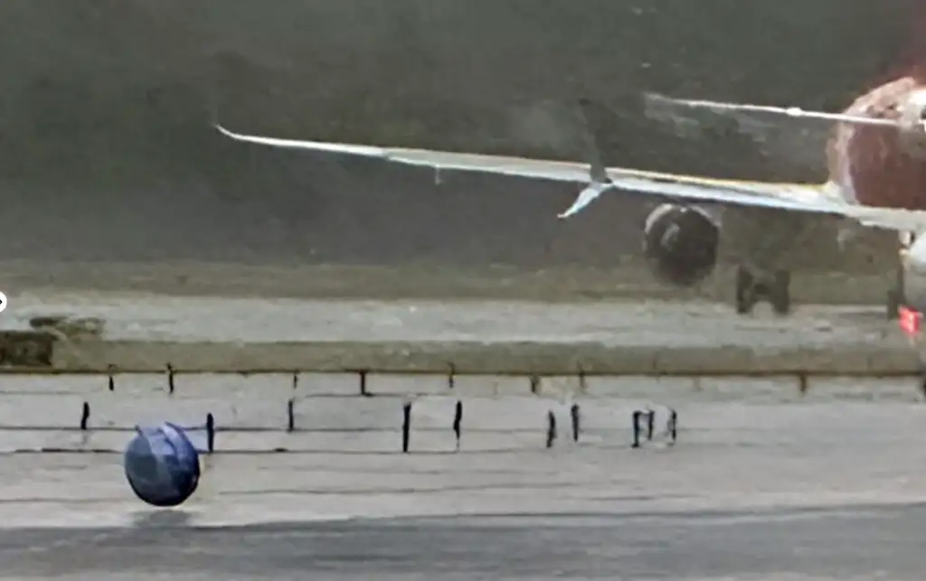 An airplane is partially visible on a tarmac, with its landing gear touching down. The scene is obscured by rain, making the surface appear wet and reflective. A row of ground lights is visible in the foreground.