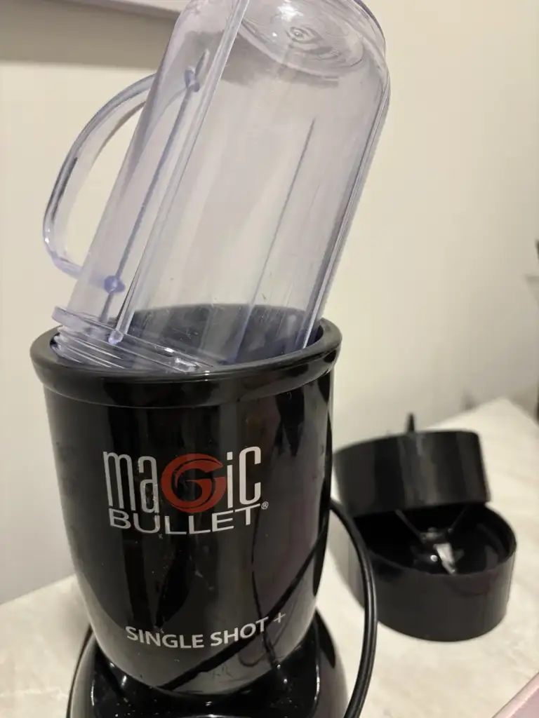 A black "Magic Bullet" blender labeled "Single Shot" with a clear blending cup inserted upside down. A black lid or base piece is visible on the side. The blender is on a light-colored surface.