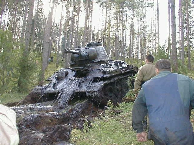 Tank being recovered in a forest with several people assisting. The ground is muddy, and the tank appears to be partially submerged. Tall trees surround the area.