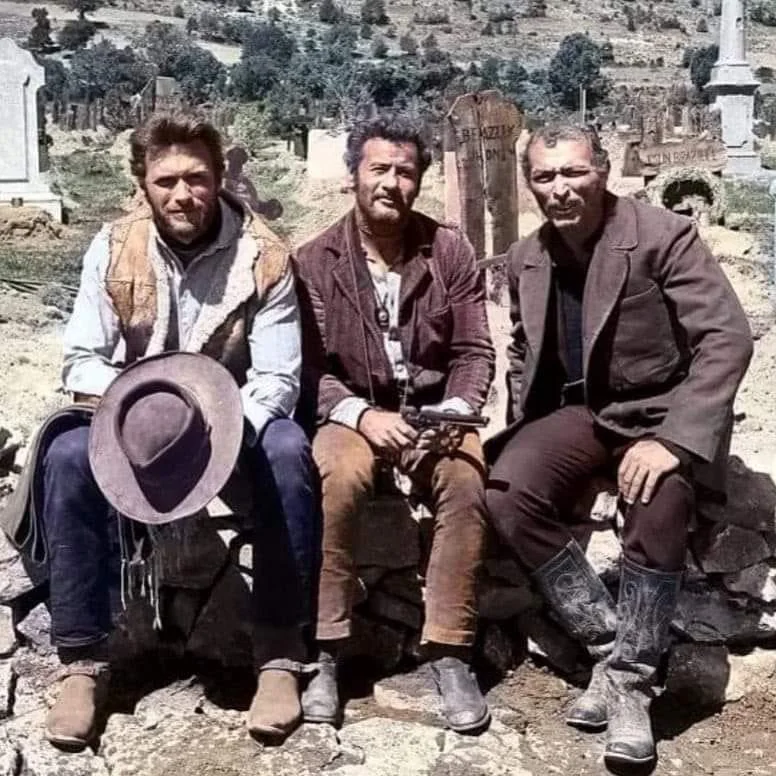 Three men in Western attire sit on a stone wall in a graveyard, with several headstones visible in the background. The man on the left holds a hat. They appear to be in a rugged, rural setting under a clear sky.