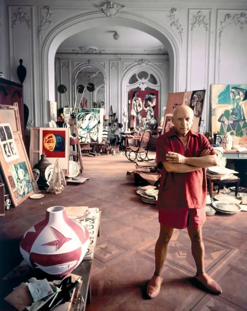 An older man stands confidently in a spacious, art-filled studio. He wears a red outfit and is surrounded by various paintings and sculptures. The room has ornate architecture, with high ceilings and wooden floors, creating an atmosphere of creativity.