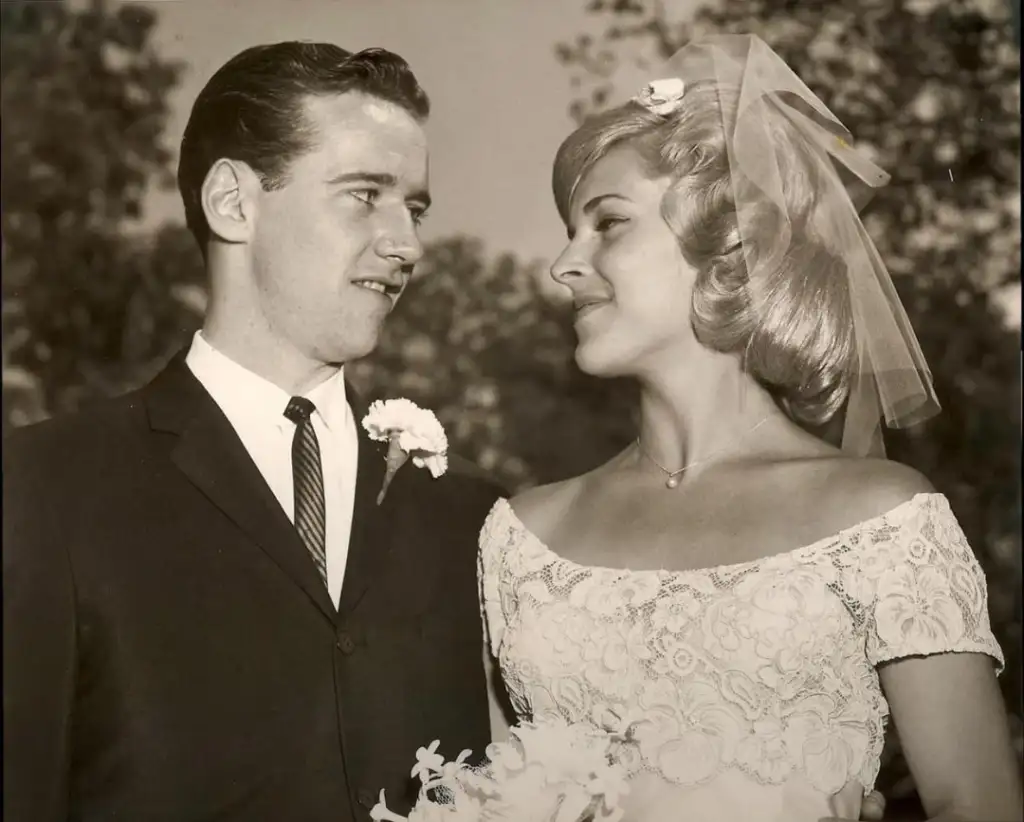 A black and white photo of a bride and groom on their wedding day. The groom wears a suit with a boutonnière, and the bride wears an off-shoulder lace gown with a veil, holding a bouquet. They gaze at each other with smiles, standing outdoors.