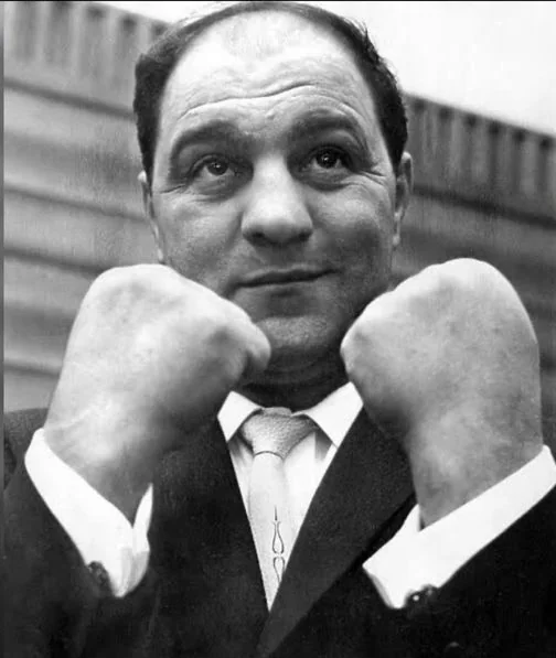 A man in a suit and tie poses with his fists raised in front of him. He has a confident expression and stands against a blurred background.