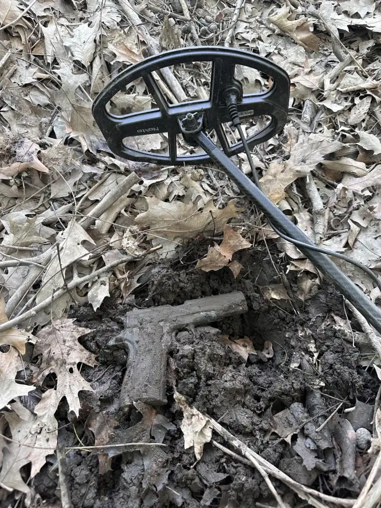 A metal detector is positioned over a partially buried, mud-covered handgun, surrounded by fallen leaves and dirt in a wooded area.