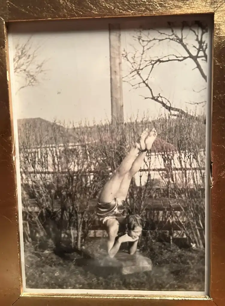 A vintage black and white photo shows a person balancing on their head in a yoga pose outdoors. They are positioned among bare bushes and a wooden fence, with leafless trees and a telephone pole in the background. The image is framed in gold.