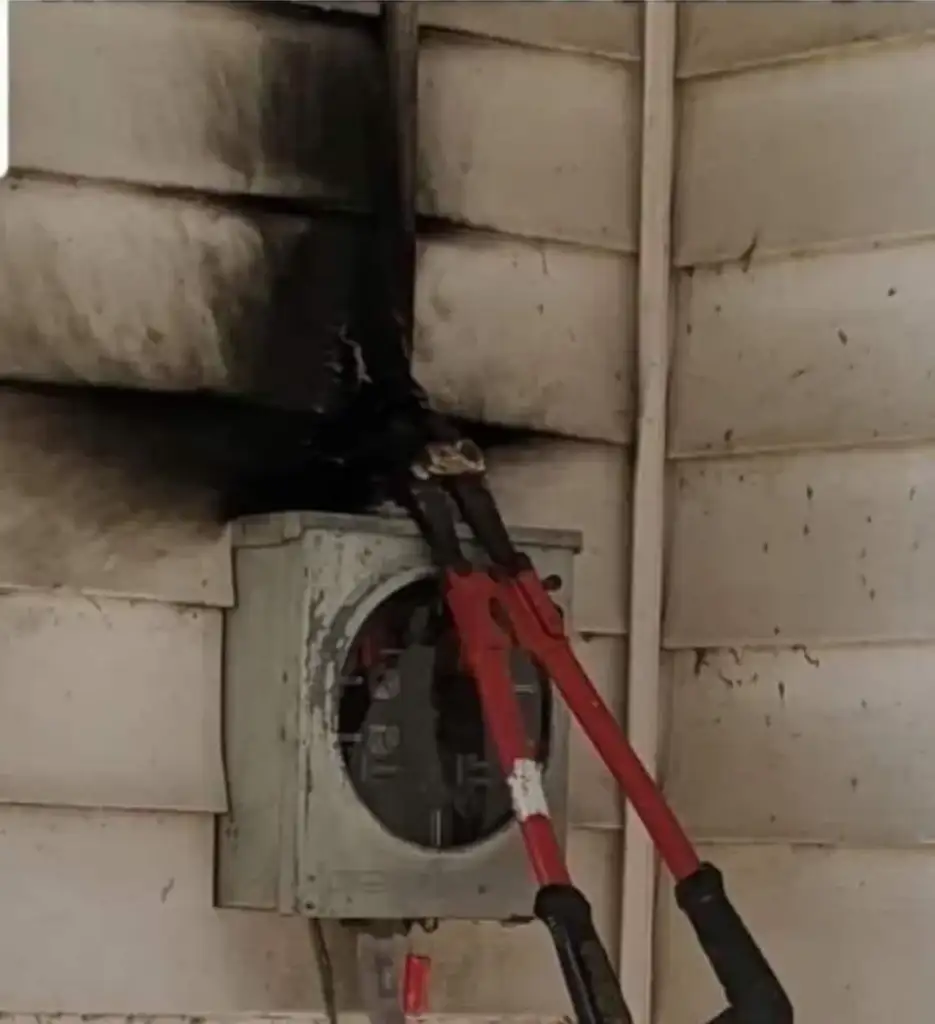 A pair of bolt cutters is wedged into a burned and blackened electrical meter box attached to the wall of a house with vinyl siding, which is stained with soot around the area.