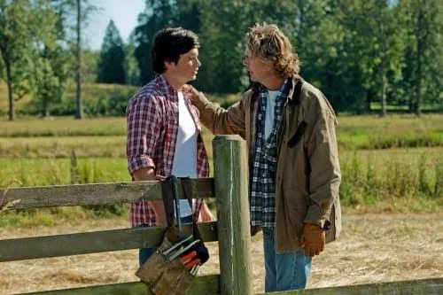 Two men are talking outdoors near a wooden fence in a rural setting. One man wears a red plaid shirt and has a tool belt hanging from the fence, while the other wears a brown jacket and gloves. A green field and trees are visible in the background.