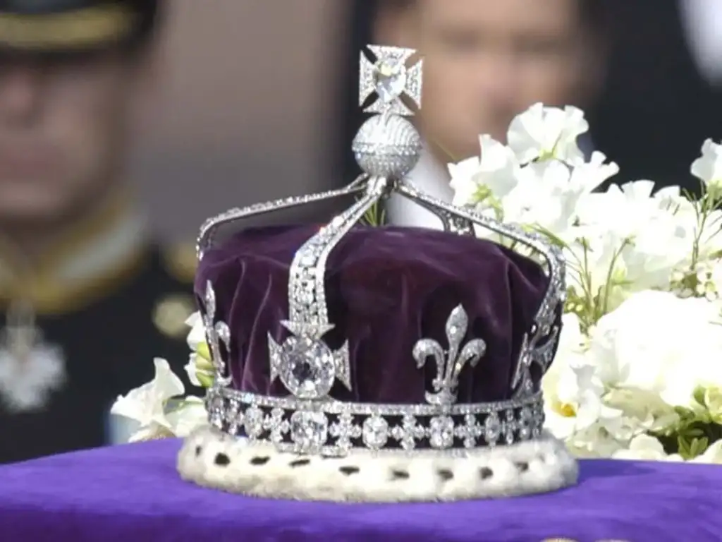 A close-up of an ornate crown placed on a rich purple cushion, adorned with diamonds and featuring intricate designs, including a prominent cross. The background is blurred, with hints of people and white flowers.