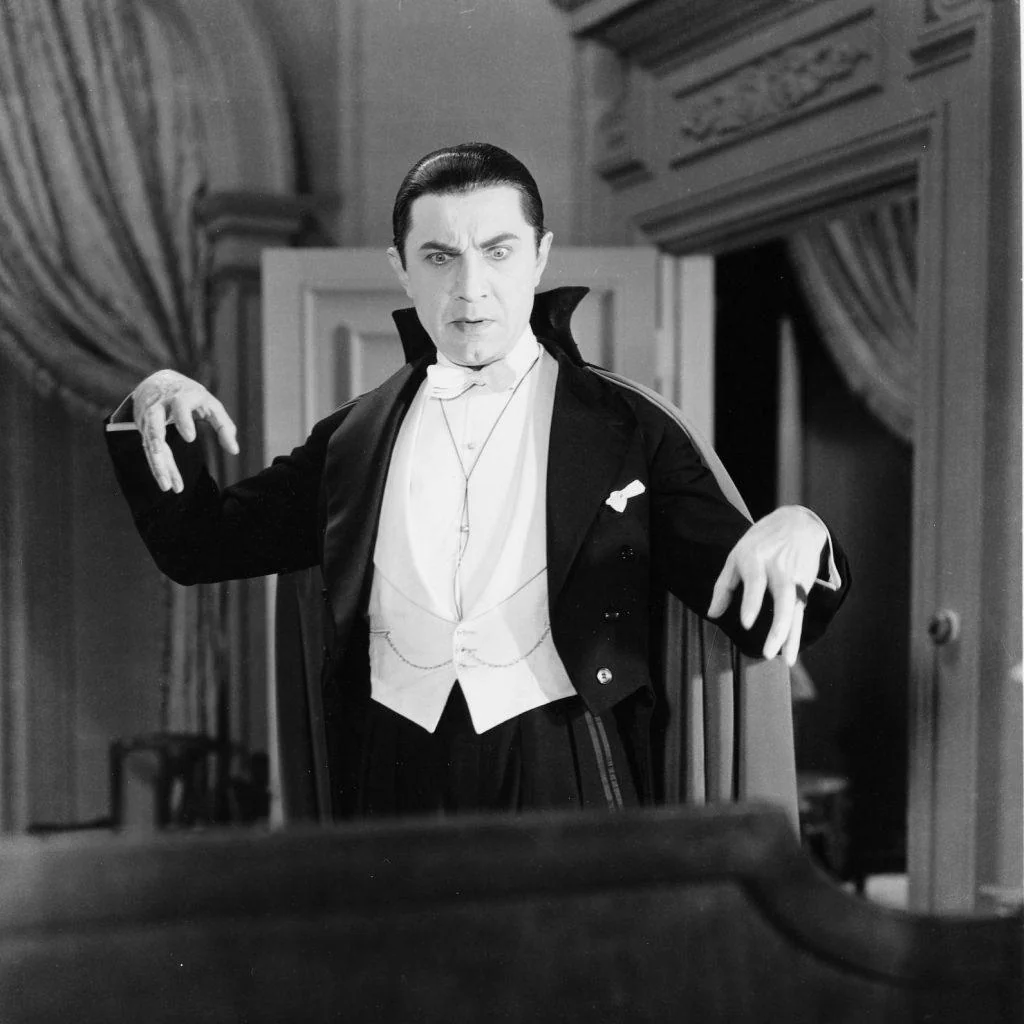 A person dressed as a classic vampire with a cape and formal attire, including a bow tie and vest, poses dramatically indoors. The background shows a room with draped curtains and a decorative doorway.