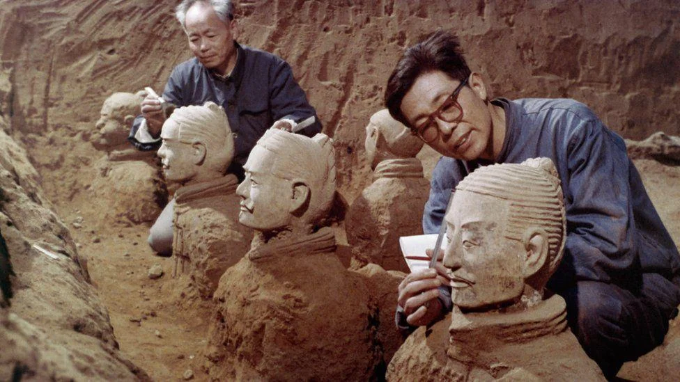 Two archaeologists carefully restore terracotta warrior statues in an excavation site. They are cleaning and inspecting the detailed faces of the figures, surrounded by earth. The site is part of the famous Terracotta Army in China.