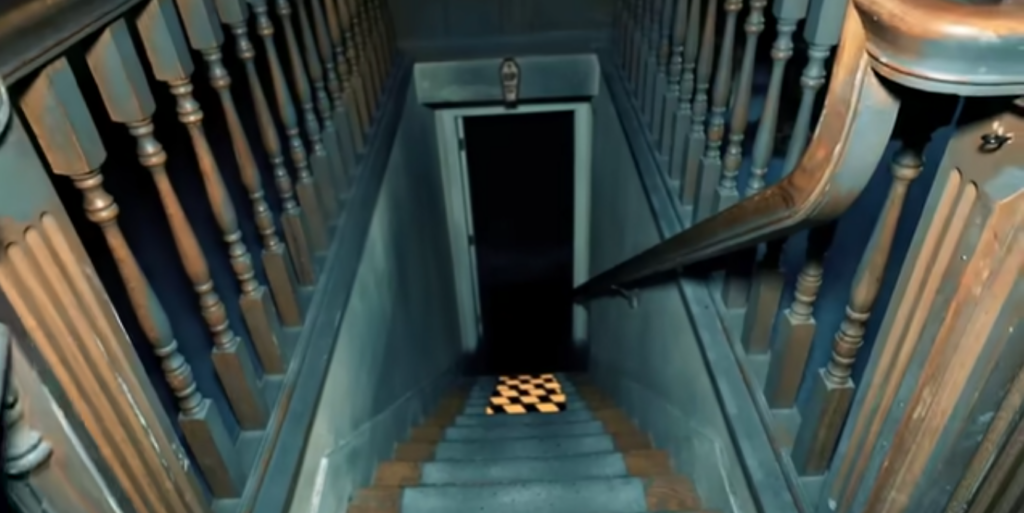 A dimly lit staircase leads down to an open door revealing darkness and a small lit checkered floor. The staircase has wooden railings and panels with an eerie, bluish tone. The perspective emphasizes depth and mystery.