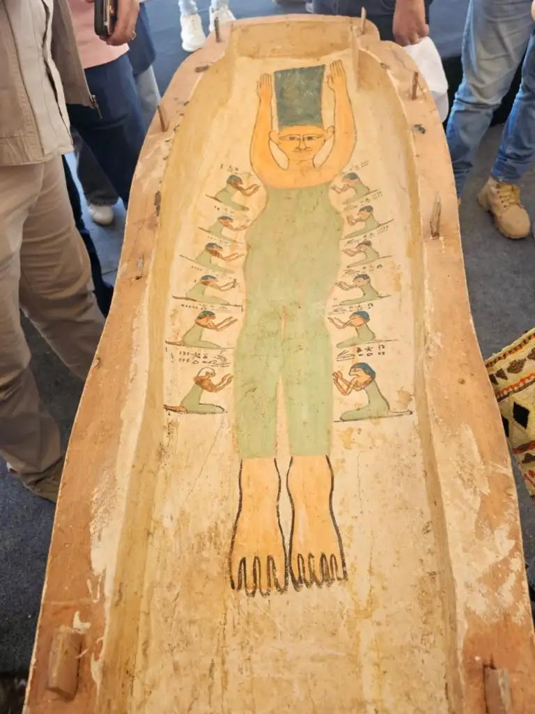 An ancient sarcophagus lid featuring a painted figure in green with arms raised. Surrounding figures appear to be offering prayers or performing rituals. A group of people is gathered around observing.