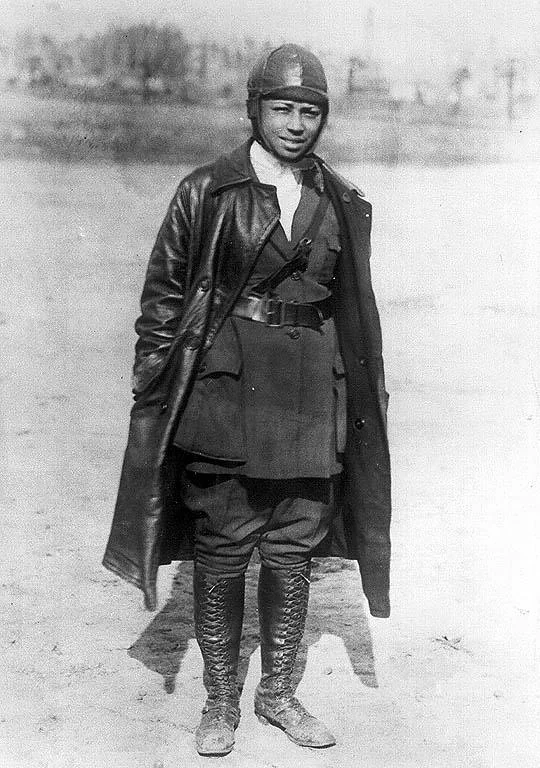 A person wearing an early 20th-century aviation outfit, including a leather helmet, goggles, long leather coat, and tall lace-up boots, stands outdoors on a dirt surface. The background is blurred, indicating an open area.