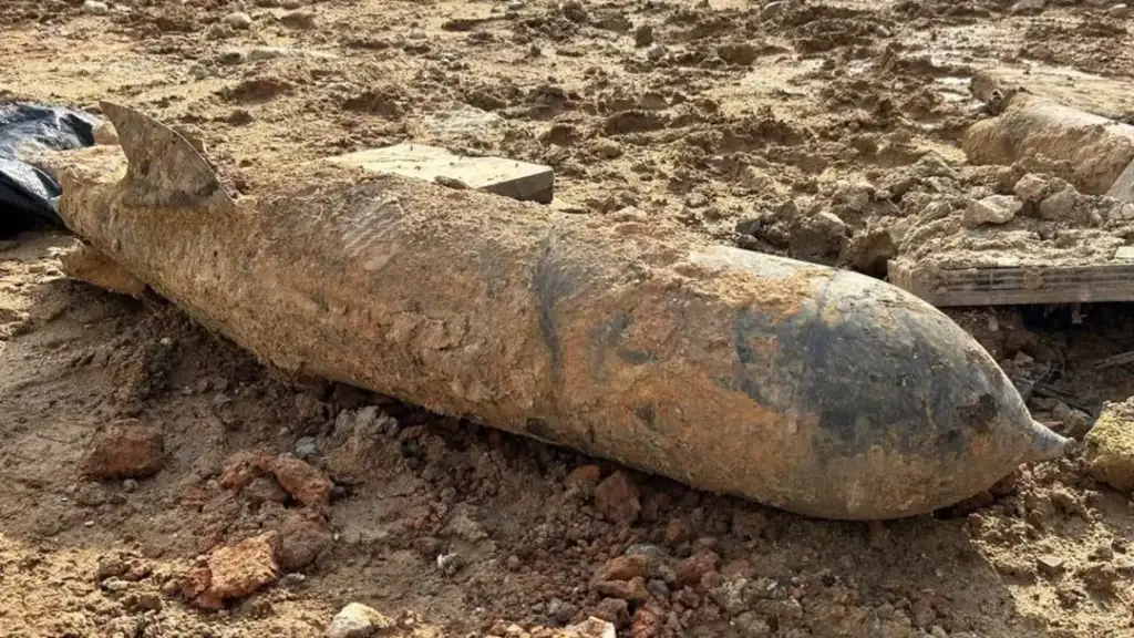 A large, rusted bomb lies partially buried in muddy, disturbed soil. The metal casing is corroded, indicating age and exposure. Surrounding debris and earth suggest recent excavation or construction work in the area.