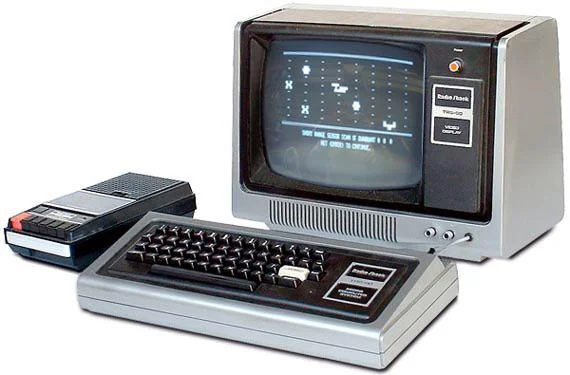 A vintage Tandy TRS-80 computer setup featuring a monochrome monitor displaying basic graphics, a tactile keyboard, and a cassette tape drive for data storage, all in gray tones.