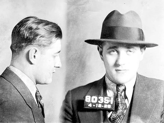 Black and white mugshots of a man wearing a suit and tie. He has short, combed hair and is also wearing a wide-brimmed hat in the front view. A sign with numbers and a date, "4-12-28," is visible in the front view.