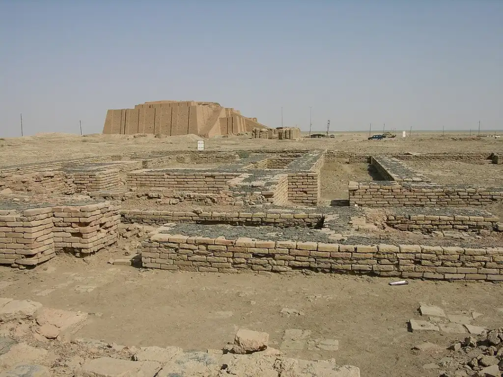 A desert landscape with ancient brick ruins. In the background, a large ziggurat structure rises. The foreground shows several lower, rectangular brick foundations, all under a clear sky.