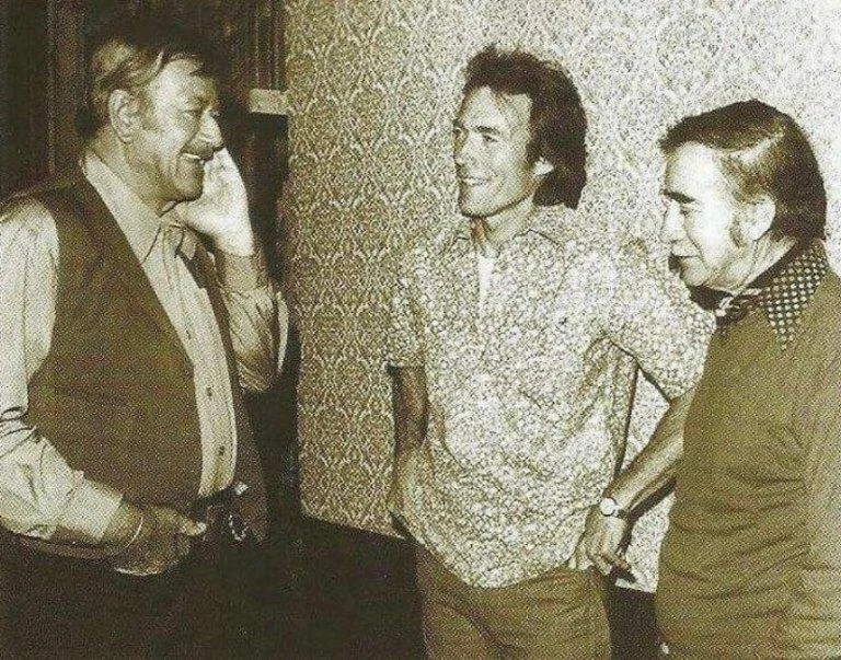 Three men stand casually in conversation in front of a patterned wall. The man on the left holds his hand to his face, the man in the middle wears a floral shirt, and the man on the right leans with his hand in his pocket. The image is sepia-toned.