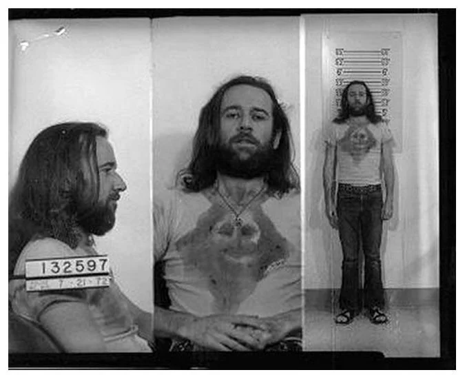 A man with long hair and a beard is shown in three black and white mugshot photos. He wears a T-shirt with a design, jeans, and sandals. The image displays front, side, and full-body views against a height chart.