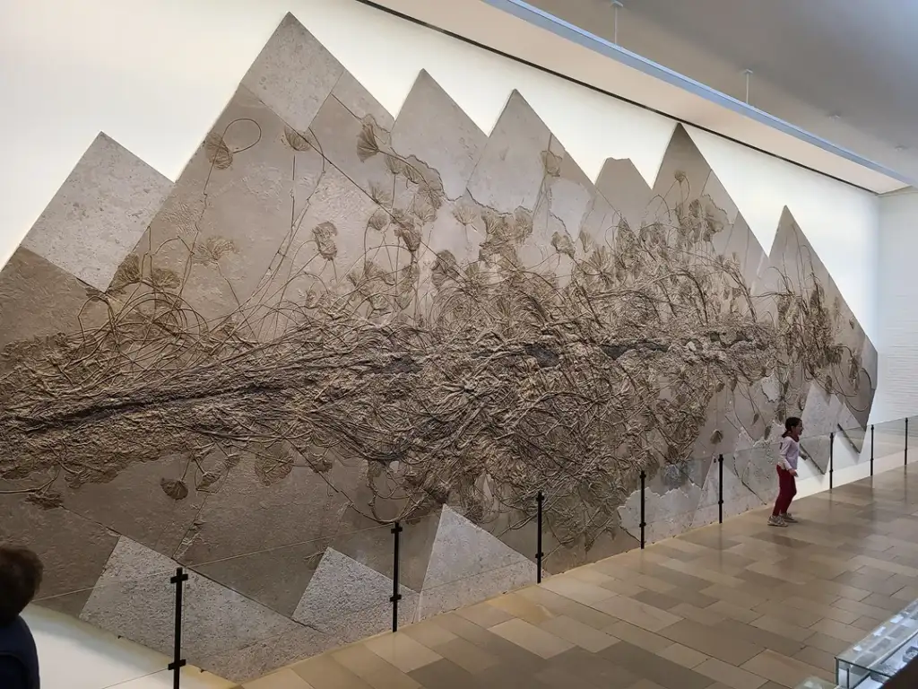 A large, intricate wall sculpture featuring geometric shapes and swirling, vine-like patterns. A child stands nearby for scale in a spacious, well-lit gallery. The floor is tiled, and the artwork is enclosed by a low barrier.