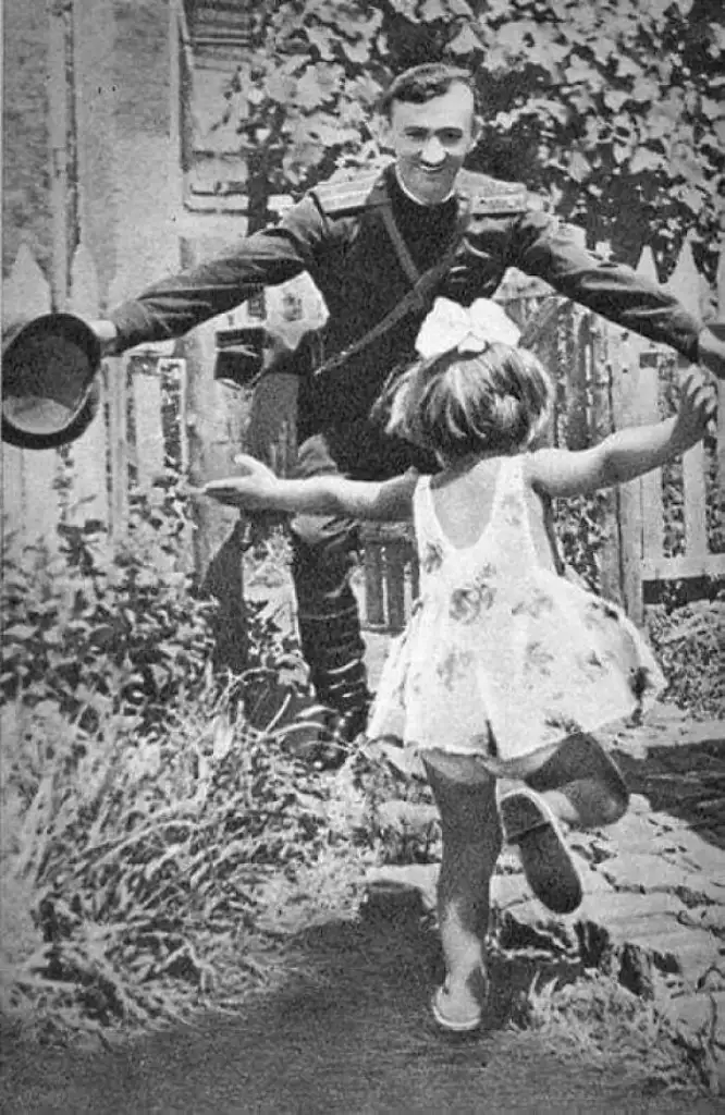 A soldier joyfully greets a little girl who is running towards him with open arms, holding a hat in one hand. They are outdoors, with a fence and foliage in the background. The girl wears a dress with a bow in her hair.