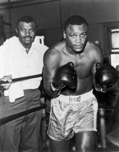 A boxer wearing Everlast shorts and gloves stands poised in a boxing ring. Next to him is a man holding a towel, possibly a coach or trainer. The scene suggests a training session or preparation for a match.