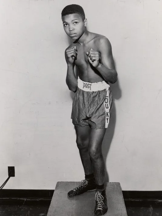 Young male boxer posing in a fighting stance. He is shirtless, wearing boxing shorts with "POKY" written on the waistband, and boxing shoes. The background is a plain wall.