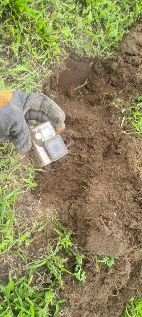 A gloved hand holds a small, dirty electronic device over a freshly dug patch of soil. Surrounding the hole is lush green grass.