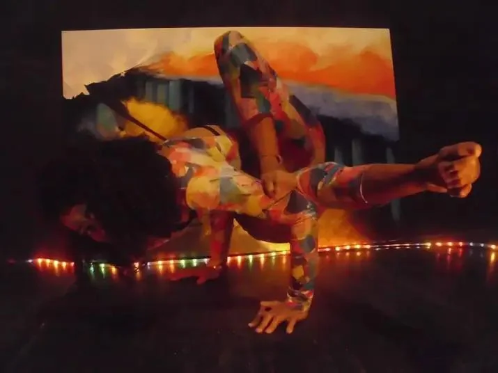 A person performs a dynamic yoga pose in a colorful, abstract bodysuit against a vibrant backdrop. The image is illuminated by a string of multicolored lights along the floor.