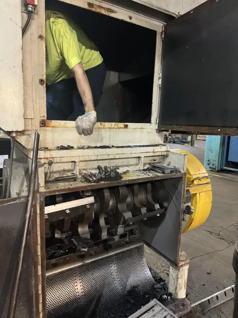 A person wearing a yellow shirt and gloves is reaching into a large industrial machine, possibly for maintenance or repair. The machine has metal components and a grated bottom section, with scattered black debris on the ground.