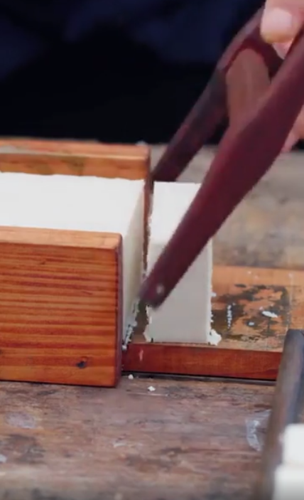 A person using a wire cutter to slice a block of tofu on a wooden board.