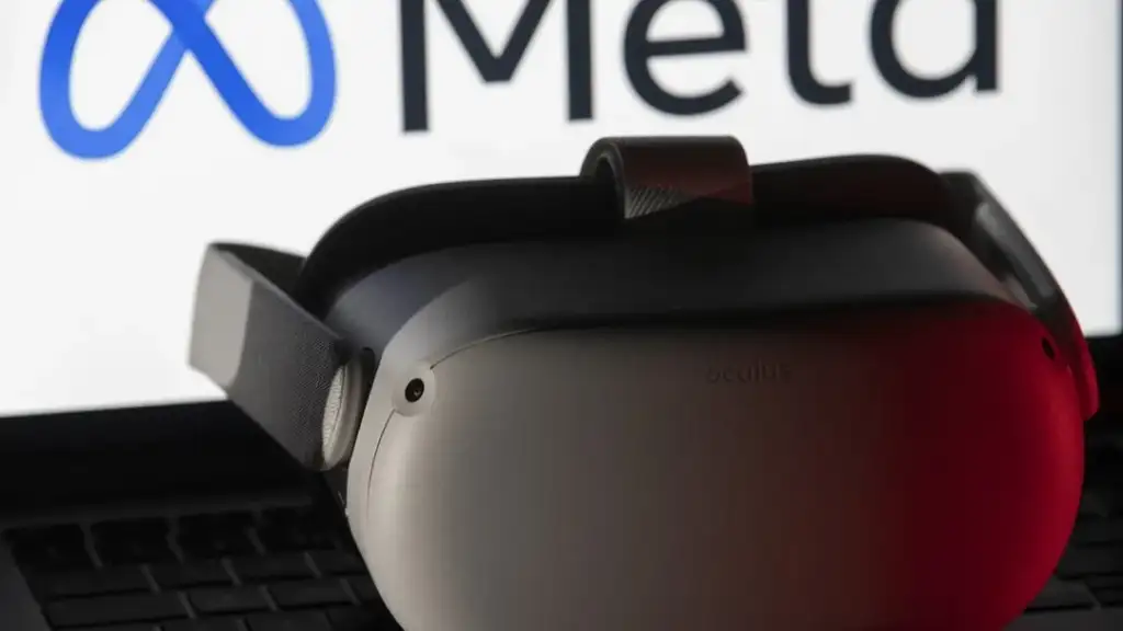 A virtual reality headset rests on a laptop, with the Meta logo visible on the screen in the background. The scene is dimly lit, highlighting the headset's contours.