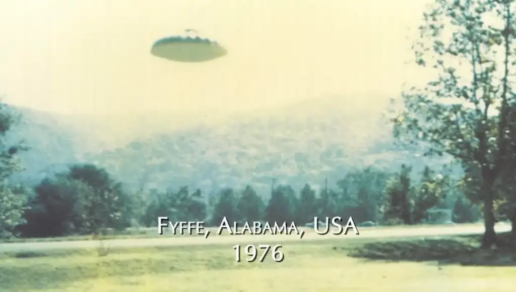 A vintage photo shows a blurred, unidentified flying object (UFO) in the sky over a rural landscape with trees and hills. The text "Fyffe, Alabama, USA, 1976" is overlaid at the bottom.