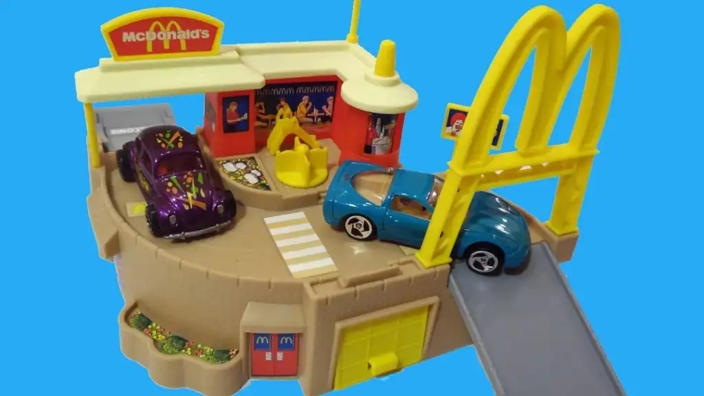 Toy McDonald's playset on a blue background, featuring a drive-thru with two toy cars: a purple one with designs and a blue one. The set includes a large arch, seating area, and small garden details.