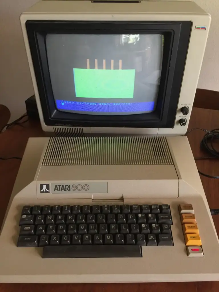 A vintage Atari 800 computer sits on a table, with a beige keyboard featuring black keys and several function keys in different colors. The monitor displays a simple graphics screen with green and blue colors.