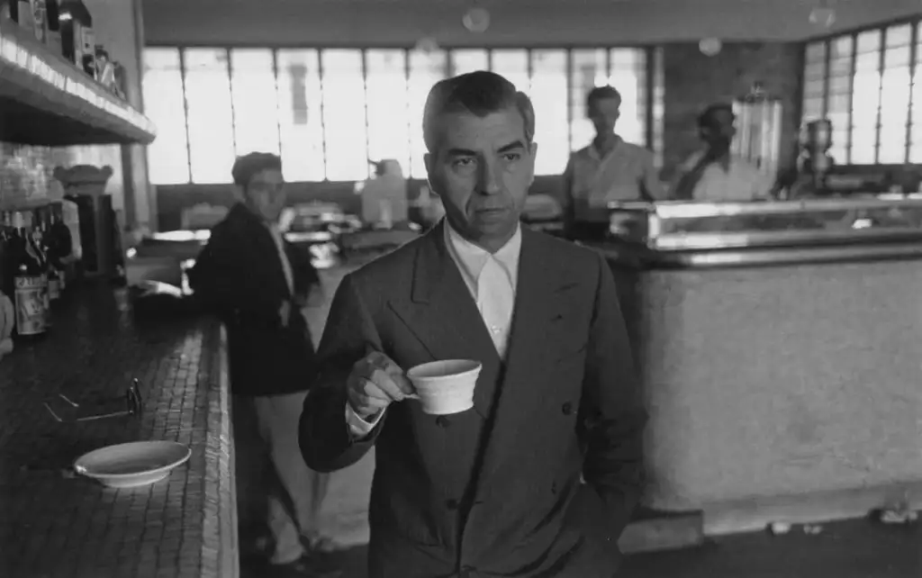 A serious man in a suit holds a coffee cup with one hand, standing at a bar counter in a café. Behind him are people in the background, partially obscured by shadows. Large windows let in natural light.