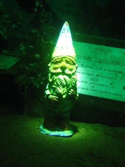 A garden gnome with a pointed hat is illuminated underwater, surrounded by faint greenery. A partially visible memorial plaque is in the background, creating an eerie atmosphere.