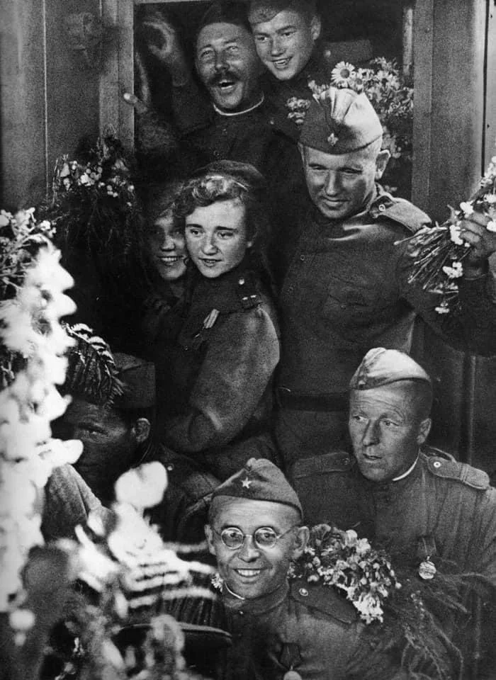 A group of soldiers in uniform are smiling and holding bouquets of flowers. They are gathered closely together, some wearing hats with stars. The mood is joyful and celebratory.