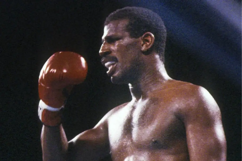 Boxer in action, shirtless, wearing red gloves, appears focused and intense, mouth slightly open. The background is dark, with a spotlight creating dramatic lighting.