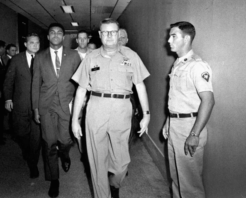 A group of men walks down a corridor. A person in a suit is flanked by others in military uniforms. The corridor is lit with overhead lights, and the scene appears formal.