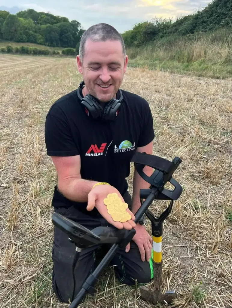 Man kneeling in a field holds a stack of golden coins in his hand. He is smiling, wearing a black shirt with logos, and has headphones around his neck. A metal detector and a small shovel are beside him. Trees are visible in the background.