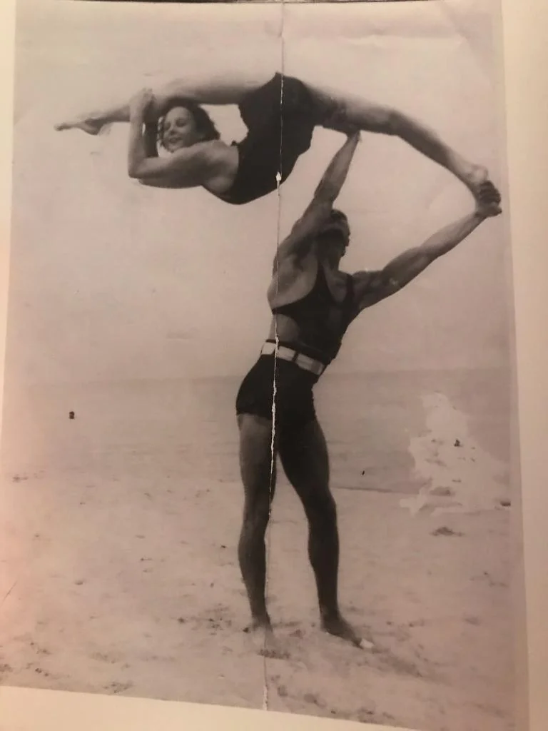 A black and white photo shows a man standing on a beach, holding a woman aloft in an acrobatic pose. She is balanced horizontally, legs outstretched. Both wear vintage-style swimsuits. The ocean is visible in the background.