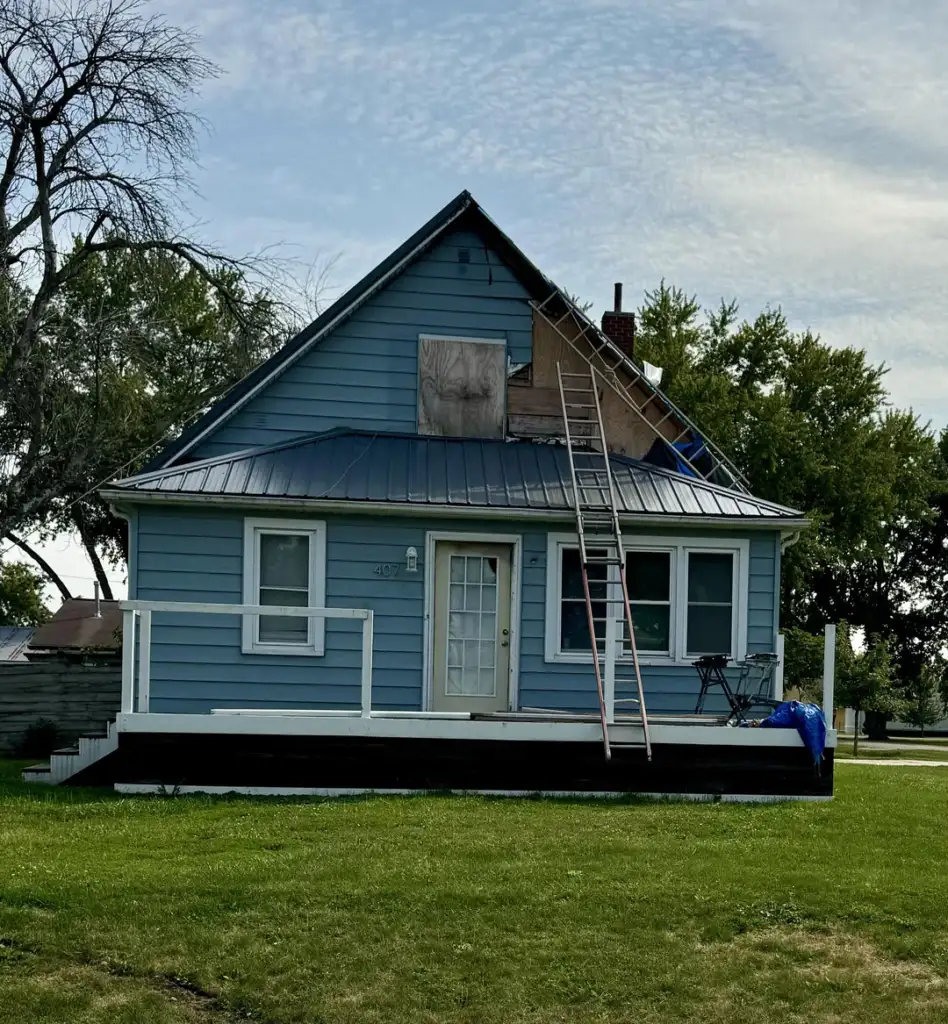 A blue house with a steep roof and a white front door is under construction. A ladder leans against the side, reaching the roof where part of the siding is removed. The house is surrounded by green grass and trees under a cloudy sky.