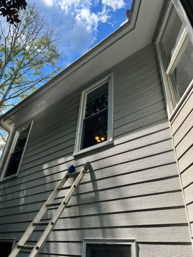 A two-story house with gray siding and white trim is shown from below. A ladder leans against the house reaching up to a second-story window. The sky is clear with a few clouds, and a tree is partially visible on the left.