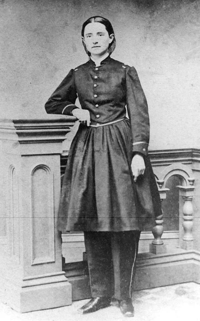 Black and white portrait of a woman in 19th-century attire, standing with one hand resting on a wooden podium. She wears a dark, long-sleeved dress with button details and a high collar, her hair pulled back neatly. The setting appears formal.