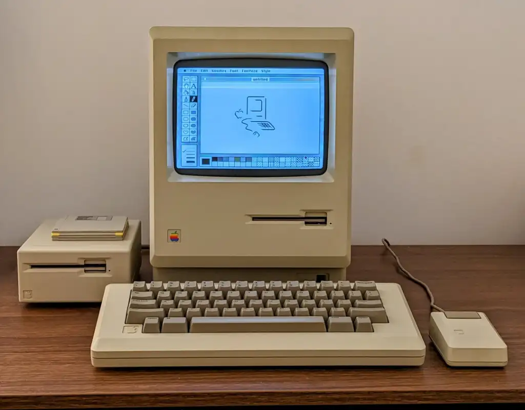 A vintage beige desktop computer from the 1980s on a wooden desk. It features a built-in monitor displaying a pixelated image, with a detached keyboard and a wired mouse. Two external disk drives are positioned to the left of the monitor.