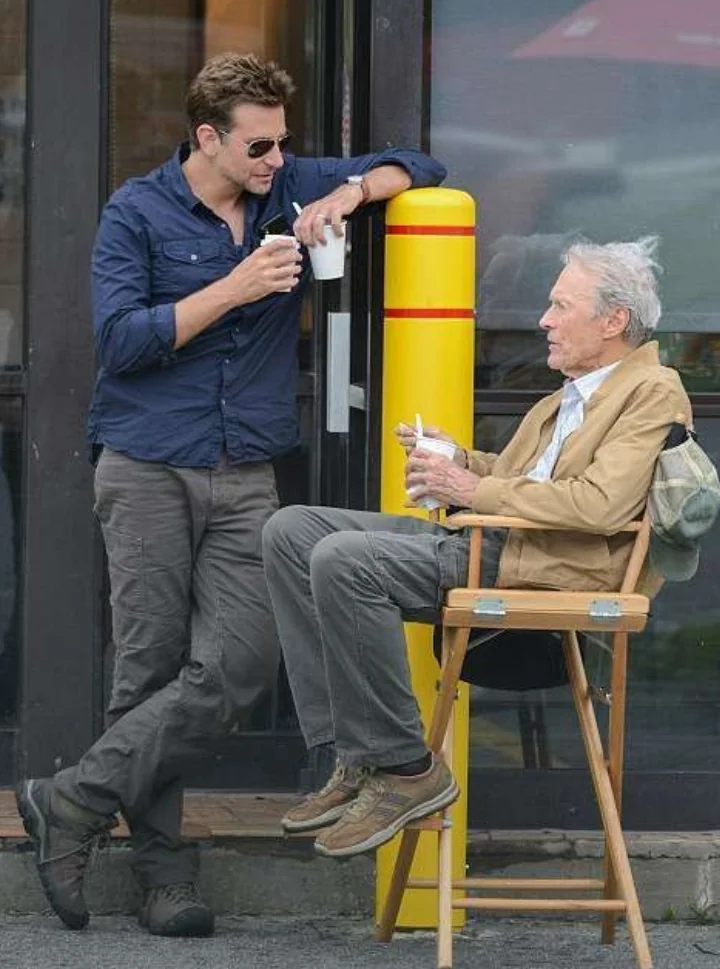 Two men are outdoors, chatting. One is seated on a wooden director's chair, wearing a beige jacket and brown shoes, holding a drink. The other leans on a yellow post, wearing sunglasses and a blue shirt, also holding a drink.