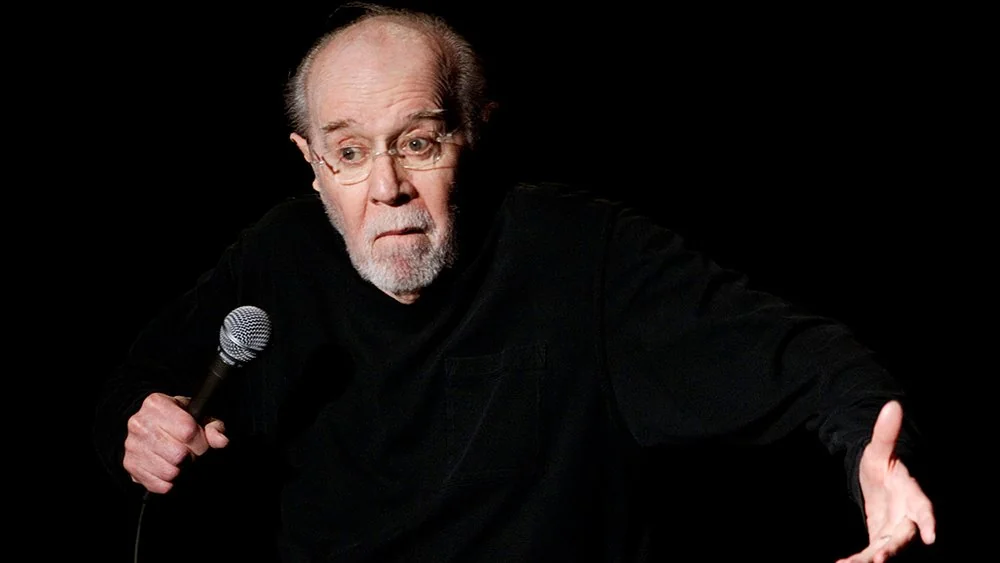 A person with a beard stands on stage holding a microphone, dressed in black, against a dark background. They have an expressive face and one hand is outstretched.