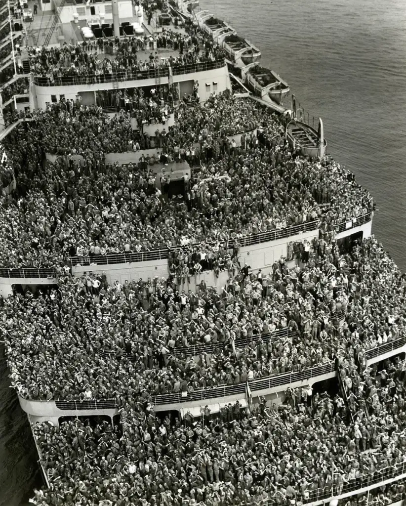 Black and white photo of a large ship crowded with people, filling the decks from front to back. The image conveys a sense of movement and anticipation as the ship is at sea, with water visible on the right.